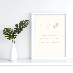 May include: A white framed wall art print featuring three small botanical illustrations and the text: learn by doing, grow by reflecting, change by adapting. Beside the frame is a white vase containing three green monstera leaves.