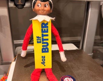 May include: A small elf doll with a red hat and suit, wearing a white collar, holding a yellow rectangular block printed with the text ELF BUTTER and .4oz in blue. The doll has dark hair and a smiling face with rosy cheeks.