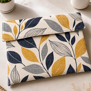 Puede incluir: Una funda rectangular de tela para portátil con cierre de solapa plegable. El exterior muestra un patrón botánico repetitivo de hojas en amarillo mostaza, azul marino oscuro y gris claro sobre un fondo texturizado de color beige claro.