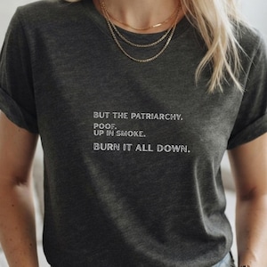 May include: A dark charcoal grey short-sleeve t-shirt featuring white text that reads: BUT THE PATRIARCHY. POOF. UP IN SMOKE. BURN IT ALL DOWN. The shirt has a crew neckline and rolled sleeves, providing a casual and straightforward appearance.