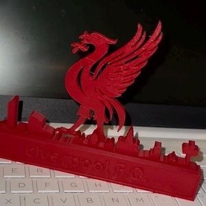 Può includere: Un oggetto decorativo rosso stampato in 3D raffigurante l'emblema dell'uccello Liver del Liverpool F.C. sopra una silhouette dello skyline. La base dell'articolo mostra il testo Liverpool F.C. in rilievo con un carattere chiaro e audace.