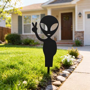 Könnte beinhalten: Ein mattschwarzer Gartenstecker aus Metall in Form einer Alien-Silhouette. Die Alien-Figur hat einen großen ovalen Kopf mit mandelförmigen Augen und einem kleinen Mund, wobei ein Arm ein Friedenszeichen zeigt. Die Figur ist auf einem Stab montiert.