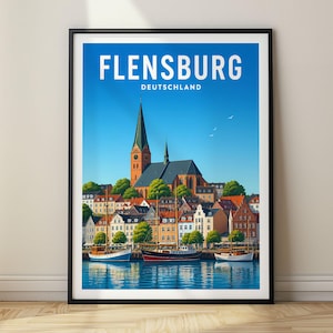 Op de afbeelding: Een ingelijste reisposter met een felblauwe lucht boven de stad Flensburg, Deutschland. De illustratie toont een hoge kerktoren, kleurrijke gebouwen aan het water met rode daken en drie zeilboten op het kalme water.