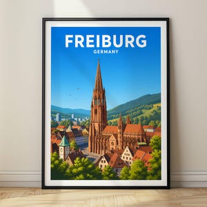 Könnte beinhalten: Ein gerahmtes Reiseplakat mit einer farbenfrohen Illustration des Freiburger Münsters in Deutschland. Das Bild zeigt den hohen Kirchturm, umliegende Gebäude mit roten Dächern und grüne Hügel unter einem strahlend blauen Himmel mit dem Text FREIBURG GERMANY.
