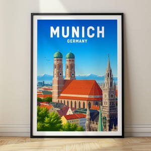 Peut inclure: Une affiche de voyage encadrée représentant le panorama de Munich, en Allemagne, avec la cathédrale Frauenkirche, des toits en tuiles rouges et des montagnes enneigées au loin sous un ciel bleu. Le texte MUNICH GERMANY est imprimé en blanc.