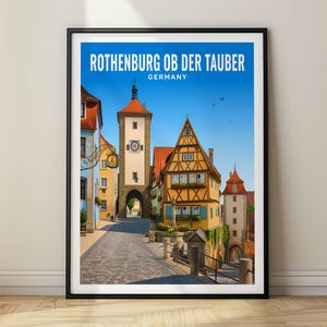 以下が含まれることがあります： ドイツのローテンブルク・オプ・デア・タウバーの街並みが描かれた額装済みのトラベルポスター。石畳の道、高い石造りの時計塔、黄色い木組みの建物が、晴れ渡った青空の下に描かれており、上部にはテキストが配置されています。