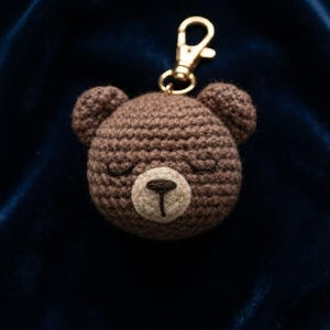 Puede incluir: Una cabeza de oso tejida a ganchillo en color marrón con un hocico beige claro, nariz y boca bordadas en marrón oscuro y ojos cerrados. La cabeza del oso está unida a un mosquetón de metal dorado para engancharla fácilmente a bolsos o llaves.