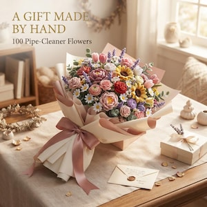 Op de afbeelding: Een boeket van 100 bloemen gemaakt van pijpenragers met zonnebloemen, rozen, tulpen en lavendel, verpakt in lichtroze en crèmekleurig papier met een bijpassend lint. De tekst A GIFT MADE BY HAND 100 Pipe-Cleaner Flowers is boven het boeket zichtbaar.
