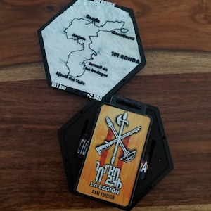Puede incluir: Dos estuches hexagonales negros. Uno contiene un mapa blanco de la ruta 101 Ronda con etiquetas de texto negro para lugares como Ronda y Setenil de las Bodegas. El otro contiene una medalla rectangular con armas cruzadas y el texto 101 KM 24h LA LEGION XXVI EDICIÓN.