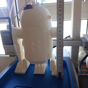 Puede incluir: Una figura de droide impresa en 3D de color blanco con cuerpo cilíndrico y cabeza abovedada, sobre una base azul con el texto STAR WARS. Una regla metálica colocada verticalmente junto a la figura muestra una altura de unos 24 cm.
