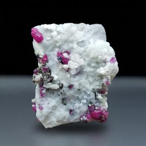 May include: A raw mineral specimen featuring a white, crystalline matrix embedded with vibrant, deep pink ruby crystals and small, dark metallic inclusions scattered across the textured, uneven surface of the stone formation.