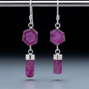May include: A pair of drop earrings featuring silver hooks and settings. Each earring displays a hexagonal magenta gemstone attached to a cylindrical, textured magenta stone piece, creating a vertical, dual-stone design with metallic silver accents.
