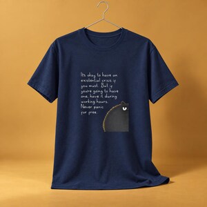 May include: A navy blue short-sleeve t-shirt featuring a graphic of a black cat peeking from the right side. The text reads: It's okay to have an existential crisis if you must. But if you're going to have one, have it during working hours. Never panic for free.