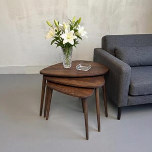 May include: A set of three nesting tables crafted from dark brown wood with a smooth finish and tapered legs. The tables feature a rounded triangular shape and are stacked together. A clear glass vase with white lilies sits on the top table.