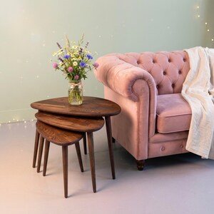 May include: A set of three dark brown wooden nesting tables with rounded, teardrop-shaped tops and tapered legs. A clear glass jar filled with a colourful bouquet of wildflowers, including daisies and lavender, sits on the largest table.