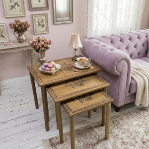May include: A set of three nested wooden tables with rectangular tops, each featuring a carved floral motif in the centre and a decorative border. The tables are arranged in a cascading formation, with a white ceramic bowl of macarons on top.