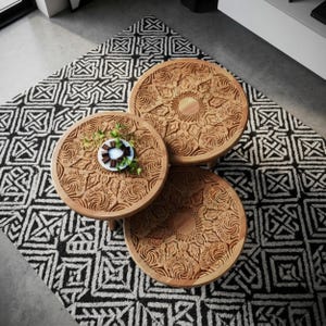 Puede incluir: Tres mesas redondas de madera con intrincados patrones tallados de estilo mandala en sus superficies. Las mesas están dispuestas en grupo, y un pequeño plato blanco con cuadrados de chocolate negro y hojas verdes descansa sobre la mesa superior.