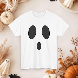 May include: A white short-sleeved t-shirt featuring a black graphic print of a ghost face. The design consists of two large vertical oval shapes representing eyes and a smaller centered oval shape representing an open mouth on the chest area.