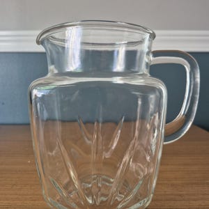 Puede incluir: Una jarra de cristal transparente con una boca ancha, un asa lateral resistente y un patrón decorativo de rayos de sol o abanico grabado en las paredes exteriores inferiores. El recipiente tiene un borde liso y una base gruesa y estable.