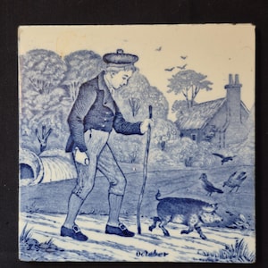 May include: A square ceramic tile featuring a blue and white transfer print of a person wearing a cap, jacket, and breeches, walking with a wooden staff. A pig walks alongside them in a rural landscape with trees, birds, and a cottage. The word October is printed at the bottom.