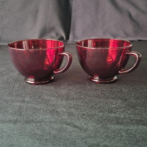 May include: Two matching teacups crafted from deep red, translucent glass. Each cup features a rounded bowl shape, a small circular base, and a curved handle on the side. The glass has a smooth, reflective surface that catches light, showing a dark crimson hue.