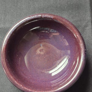May include: A small, round ceramic bowl with a glossy, deep purple glaze. The interior surface features a smooth, reflective finish with subtle variations in color intensity, while the thick, rounded rim shows a slightly lighter purple hue.