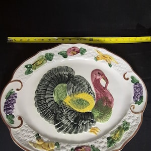 Puede incluir: Una bandeja de servicio ovalada de cerámica con un pavo en relieve en el centro, con plumas negras, verdes y amarillas y cuello rosa. El borde tiene una textura de mimbre blanca con uvas, verduras y flores pintadas. Una cinta métrica amarilla marca 53 cm.