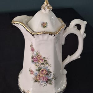May include: A white porcelain coffee pot featuring a floral pattern of pink and yellow roses with green leaves. The pot has a scalloped rim, a decorative handle, and a lid with a gold-painted finial. Gold trim accents the edges and base.