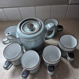 以下が含まれることがあります： 蓋付きのティーポットと6つのマグカップで構成された陶器のティーセット。各ピースはライトブルーグレーの釉薬で仕上げられ、縁とハンドルにはダークブラウンのトリムが施されています。マグカップとティーポットの内側はすべて白色です。
