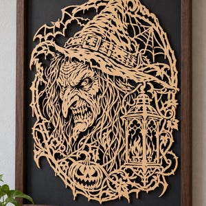May include: A framed laser-cut wood wall art piece featuring a detailed witch wearing a pointed hat, surrounded by intricate spiderwebs, thorny vines, a jack-o'-lantern, and a gothic lantern. The light-colored wood cutout is set against a black background.