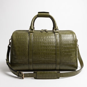 Puede incluir: Una bolsa de viaje de color verde oliva con un patrón texturizado de piel de cocodrilo. El bolso incluye dos asas superiores, una correa de hombro desmontable con una sección acolchada y herrajes de metal en tono latón con cremallera.