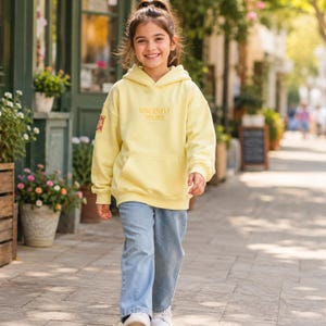 May include: A pale yellow hooded sweatshirt featuring the text SINCERELY YOURS embroidered in a matching yellow thread on the chest, a front kangaroo pocket, and a small rectangular patch on the right sleeve, paired with light blue denim jeans.