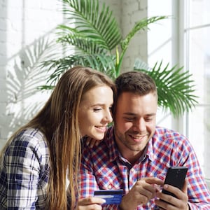 Op de afbeelding: Een zwarte smartphone en een blauwe creditcard vastgehouden door twee personen in geruite overhemden. De smartphone heeft een donker scherm en de creditcard toont een zichtbare magneetstrip en chip in een heldere, zonnige omgeving.