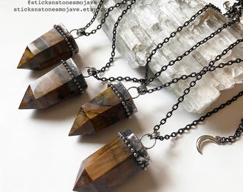 May include: Four gold and brown tiger's eye crystal point pendants on black chain necklaces. Each pendant has a silver bezel setting with a beaded edge. The necklaces are displayed on a white selenite slab. © since 2012 @sticksnstonesmojave sticksnstonesmojave.etsy.com