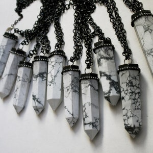 May include: A collection of white howlite crystal pendants with black metal accents hanging from black chains. The pendants are all in the shape of a long, pointed crystal.
