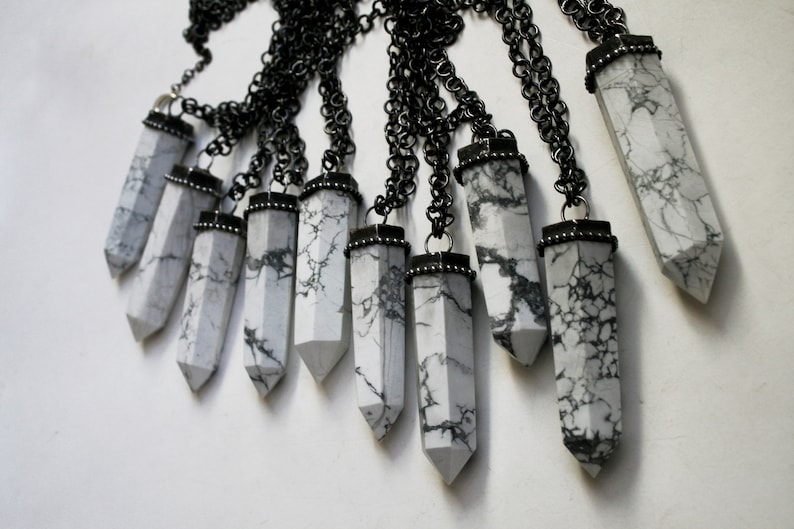 May include: A collection of white howlite crystal pendants with black metal accents hanging from black chains. The pendants are all in the shape of a long, pointed crystal.