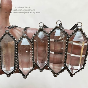 May include: Five clear, double-terminated quartz crystal pendants set in dark, beaded metal frames with attached chains. The text "© since 2012 @sticksnstonesmojave sticksnstonesmojave.etsy.com" is visible in the upper left corner of the image.