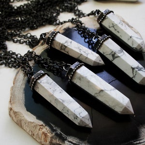 May include: Five white howlite crystal pendants with silver accents hang from black chains on a dark brown wooden surface.