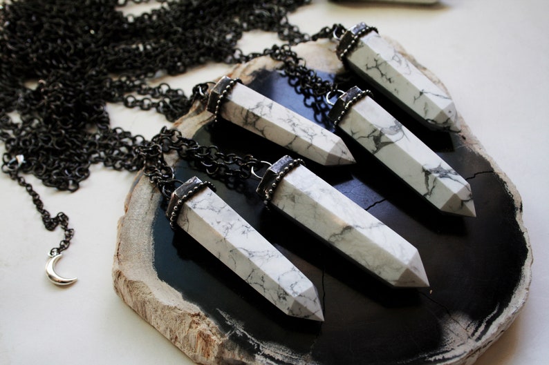 May include: Five white howlite crystal pendants with silver accents hang from black chains on a dark brown wooden surface.