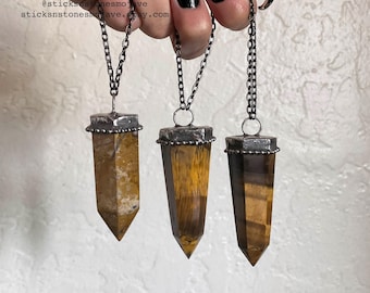 May include: Three gold and brown tiger's eye crystal point pendants on silver chains. Each pendant has a silver bezel setting with a textured edge.