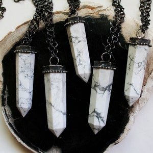 May include: Five silver necklaces with white howlite stone pendants in the shape of a long, pointed crystal. The necklaces are displayed on a dark brown wooden surface.