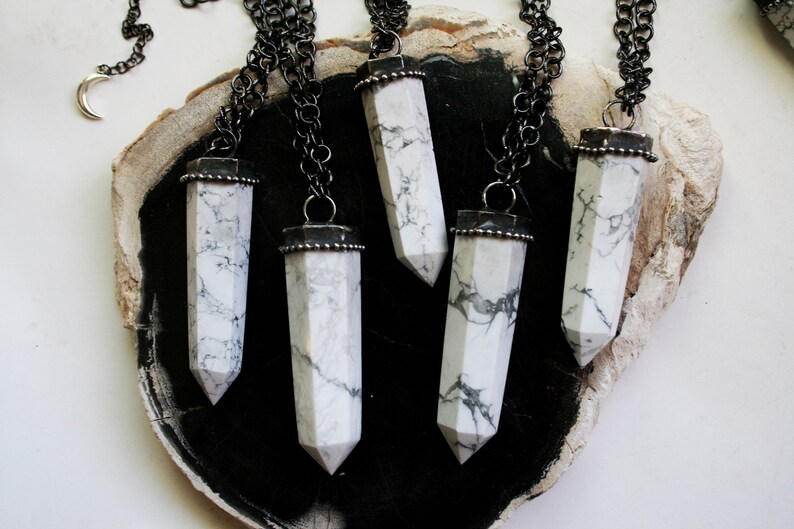 May include: Five silver necklaces with white howlite stone pendants in the shape of a long, pointed crystal. The necklaces are displayed on a dark brown wooden surface.