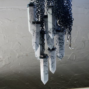 May include: A collection of white, marbled, crystal-shaped pendants hang from a black chain. The pendants are all different sizes and shapes, and they are connected to the chain by silver rings.