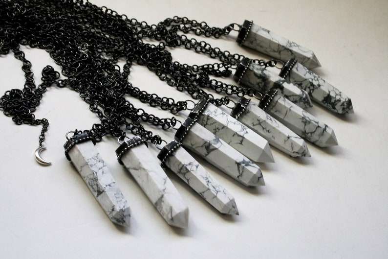 May include: A collection of white howlite crystal point pendants on black chains. The pendants are all different sizes and shapes, and they are all strung on black chains. The chains are all different lengths, and they are all hanging from a white surface.