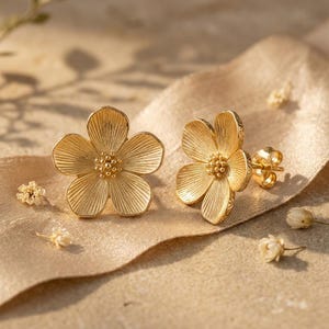 Peut inclure: Une paire de boucles d'oreilles à clous dorées en forme de fleurs à cinq pétales. Chaque boucle présente une surface nervurée et texturée sur les pétales ainsi qu'un groupe de petites sphères en relief au centre, avec une tige et un fermoir.
