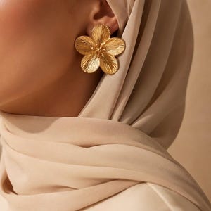 Peut inclure: Une boucle d'oreille en métal doré en forme de fleur à cinq pétales avec une finition brossée et texturée sur les pétales et un petit groupe de perles au centre. La boucle d'oreille est portée au lobe, partiellement encadrée par un foulard beige.