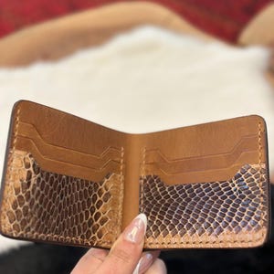 Könnte beinhalten: Ein Bifold-Portemonnaie mit einem beigefarbenen Lederinnenraum und zwei horizontalen Kartenfächern auf jeder Seite. Der untere Teil jedes Kartenfachs ist mit einem strukturierten, braun-beigen Schlangenhautmuster versehen. Sichtbare Nähte.