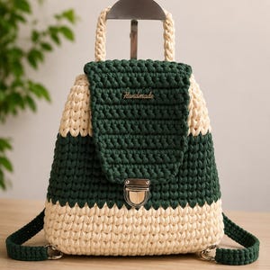 Può includere: Uno zaino all'uncinetto con strisce orizzontali alternate di filato color crema e verde scuro. La borsa include un manico superiore, una chiusura a patta con fibbia in metallo argentato e una piccola targhetta metallica con la parola Handmade.