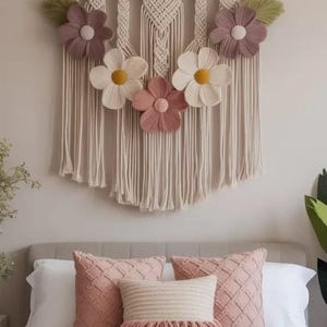 Könnte beinhalten: Ein Makramee-Wandbehang an einem Holzast mit cremefarbenen geknoteten Schnüren, fünf Stoffblumen in Mauve, Creme und Rosa mit gelben Mitten sowie zwei grünen Blattakzenten, ergänzt durch drei dekorative Kissen auf einem Bett.