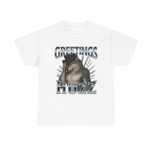 May include: A white short-sleeved t-shirt featuring a graphic of a wolf wearing a black top hat. The wolf is tipping its hat with one paw. Above the wolf is the text GREETINGS in a metallic font, and below is the text HUZZ in a metallic font.